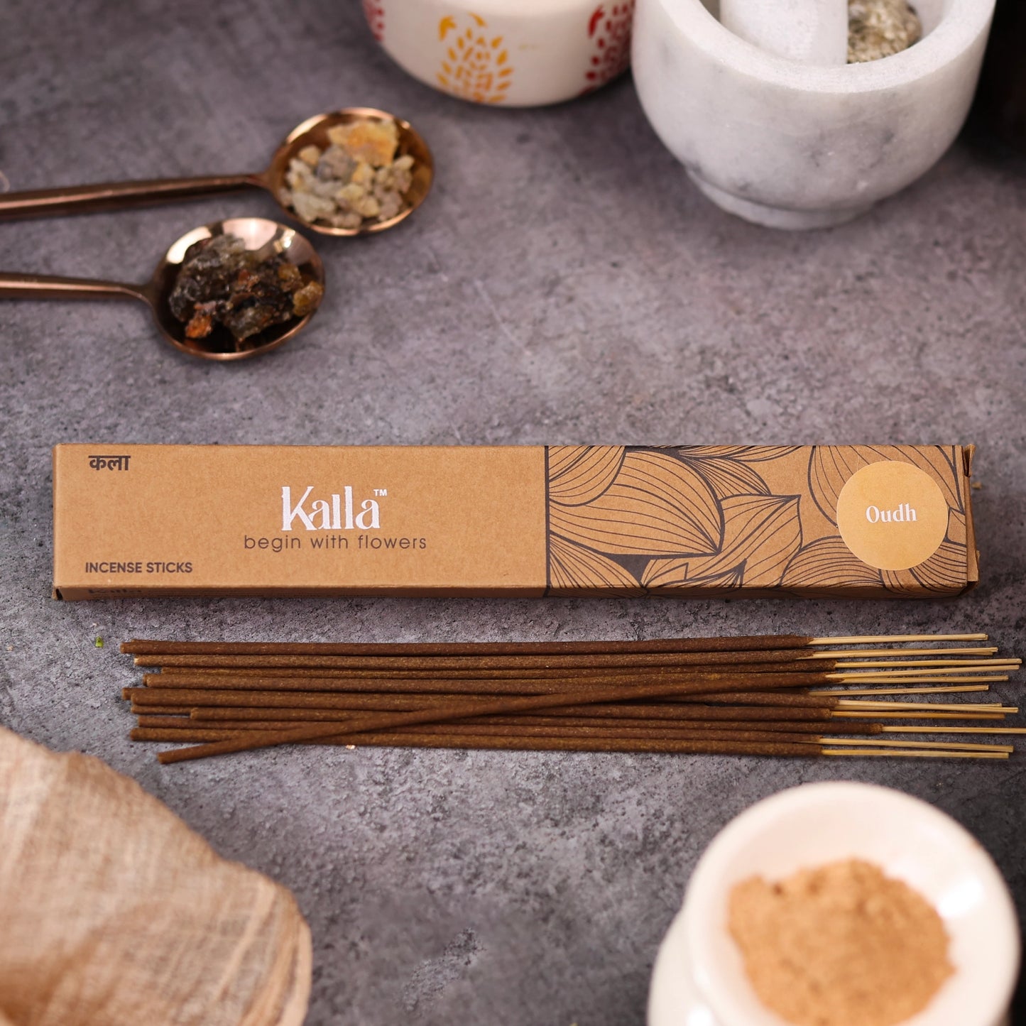Kalla Oudh incense sticks placed on a stone surface alongside the product's eco-friendly packaging. Decorative elements like brass spoons with resin, a marble mortar and pestle, and other natural ingredients surround the setup, evoking a sense of calm and luxury.