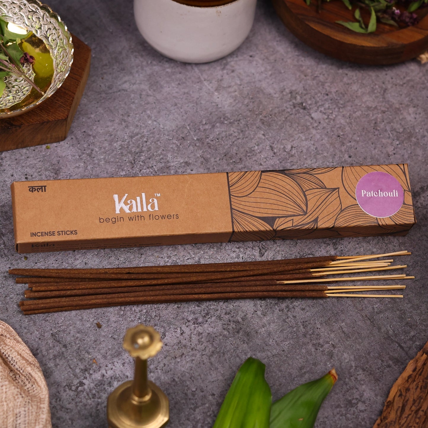 Kalla Patchouli Incense Sticks โ eco-conscious brown box with botanical line art and a purple label marked โPatchouli,โ placed on a textured stone surface alongside neatly arranged incense sticks and natural decorative elements for a calming, earthy aesthetic.