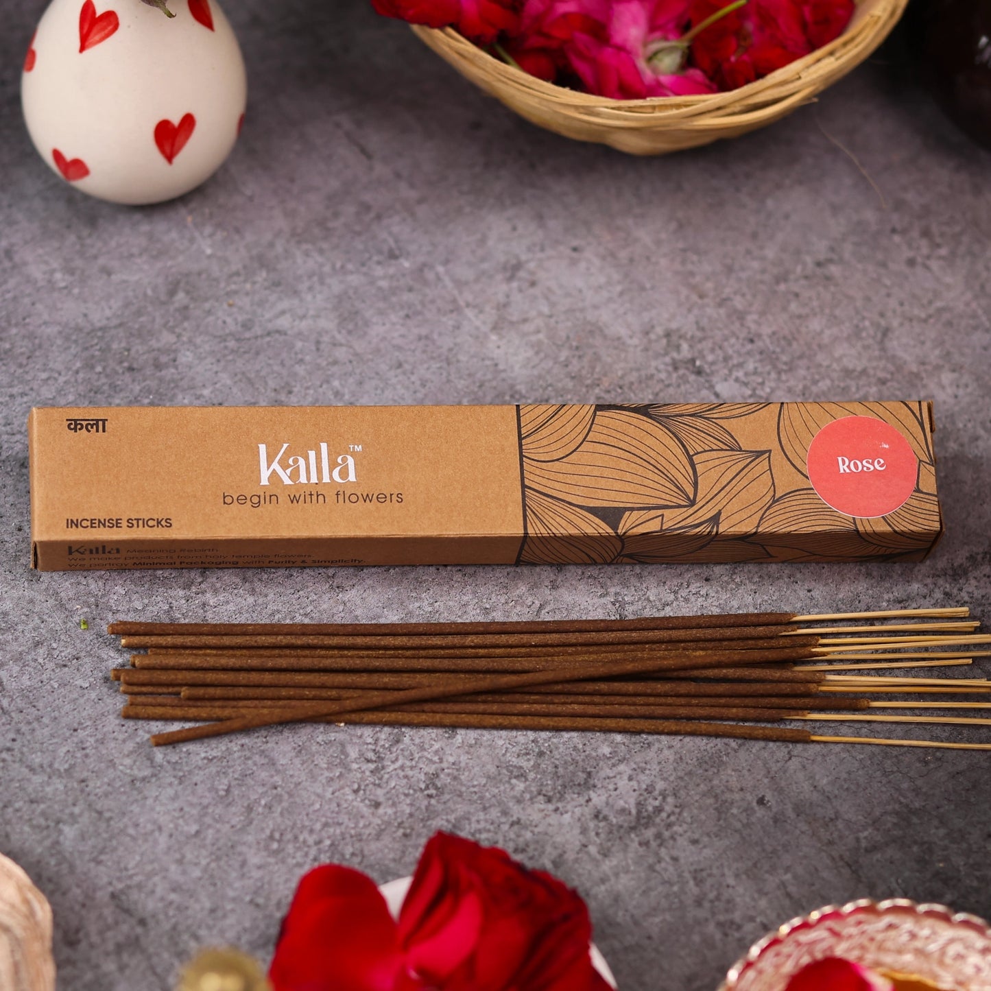 Kalla Rose Incense Sticks – natural brown packaging with elegant floral line patterns and a red label marked “Rose,” displayed on a grey stone surface with incense sticks spread out, surrounded by red rose petals and decorative elements for a calming, aromatic setting.