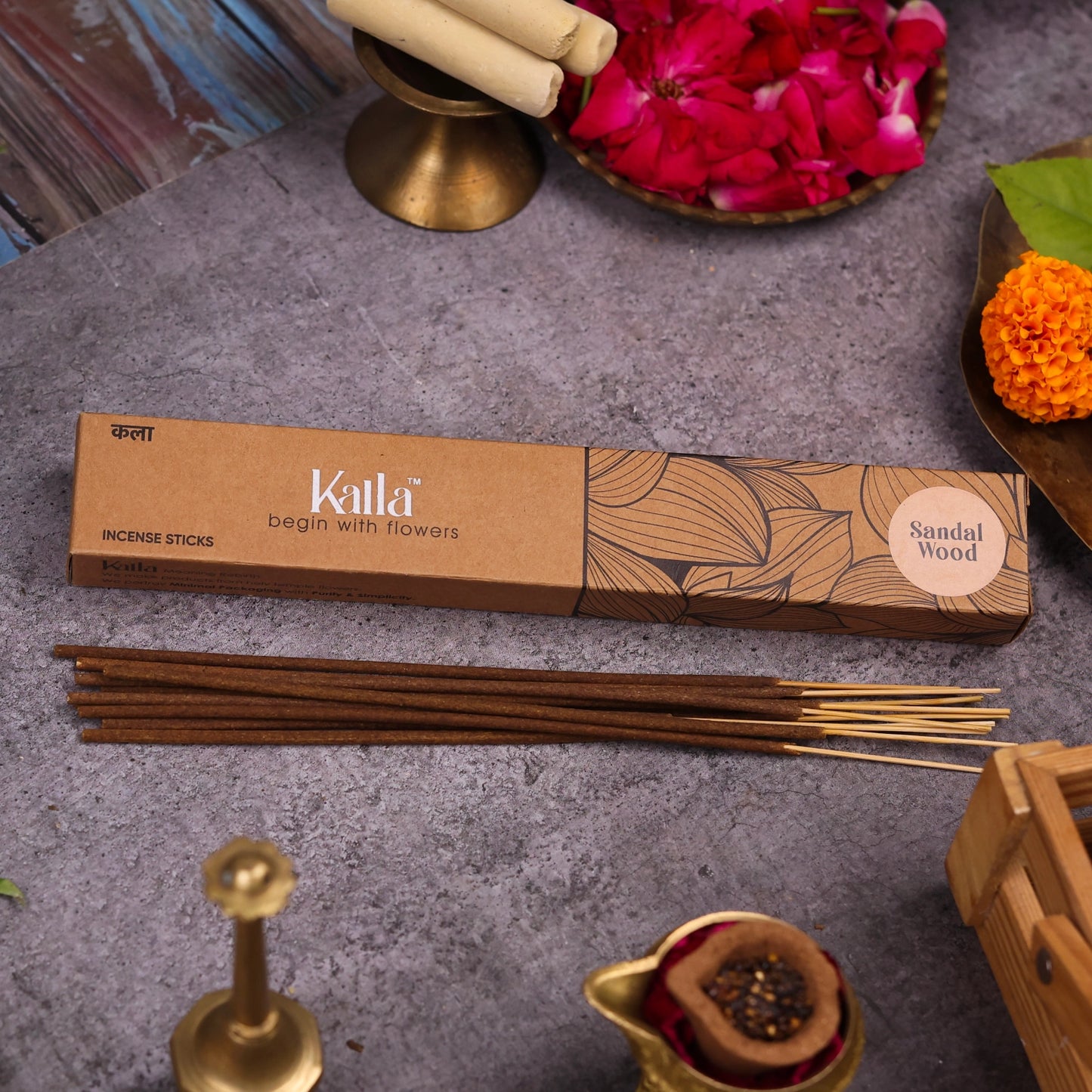 Kalla Sandalwood Incense Sticks โ a brown eco-friendly box with floral illustrations, labeled โbegin with flowersโ and โSandal Wood,โ displayed on a rustic stone background with incense sticks, rose petals, marigold flower, brass incense holder, and traditional decor elements.