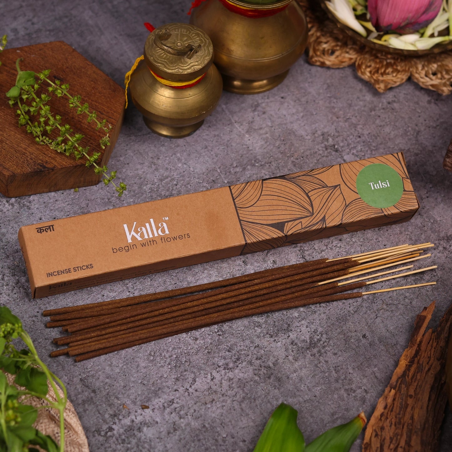 Kalla Tulsi Incense Sticks – eco-friendly brown packaging with intricate floral line art and a green label reading “Tulsi,” displayed on a grey stone surface with incense sticks laid out, surrounded by traditional brass vessels, tulsi leaves, and a serene ritual setting.