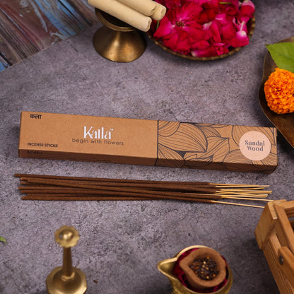 Kalla Sandalwood Incense Sticks – a brown eco-friendly box with floral illustrations, labeled “begin with flowers” and “Sandal Wood,” displayed on a rustic stone background with incense sticks, rose petals, marigold flower, brass incense holder, and traditional decor elements.